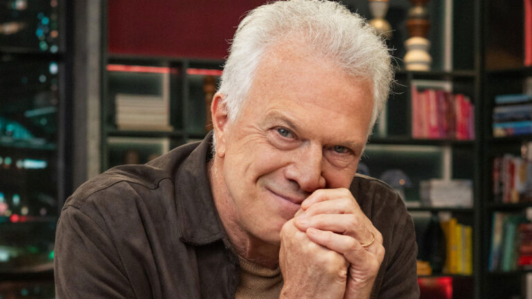 Pedro Bial sorrindo com as mãos entrelaçadas próximo ao rosto durante gravação do talk show Conversa com Bial nos estúdios da Globo