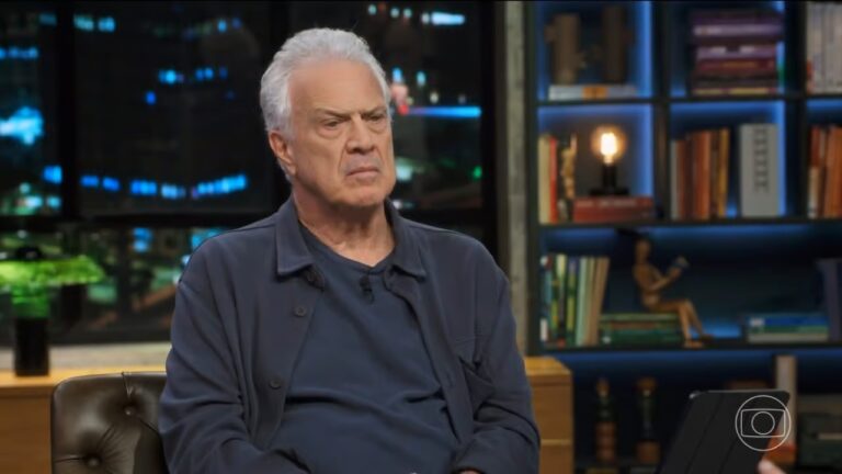 Homem de expressão séria está sentado em um estúdio de entrevistas, com estante iluminada ao fundo contendo livros e objetos decorativos.