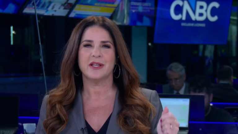 Apresentadora de telejornal aparece em estúdio moderno, em plano médio, falando diretamente para a câmera. Ela tem cabelos castanhos longos e ondulados, usa blazer cinza, blusa escura e brincos de argola. Sua expressão é séria e profissional, com uma das mãos levemente levantada em gesto de ênfase. Ao fundo, há telas, computadores e o logotipo “CNBC” em destaque, indicando um ambiente de redação de notícias financeiras.