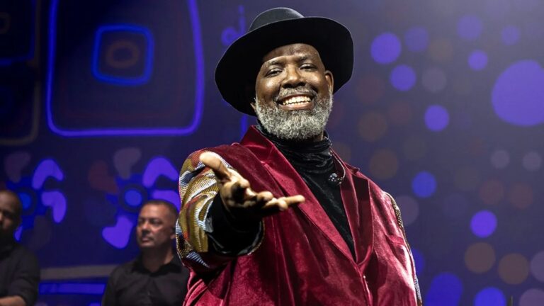 Homem sorridente em um palco, usando chapéu preto e um casaco de veludo vinho sobre roupa escura. Ele tem barba grisalha e estende uma das mãos em direção ao público, em um gesto acolhedor. Ao fundo, há iluminação de palco com tons azulados e pontos de luz desfocados, além de outro músico parcialmente visível, criando um clima de show ao vivo.