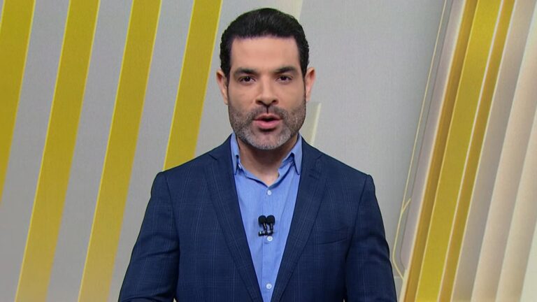A imagem mostra Darlisson Dutra vestindo terno azul-escuro e camisa azul-clara, com microfones de lapela presos à roupa. Ele está diante de um fundo com faixas diagonais amarelas e brancas, no cenário do jornal Primeiro Impacto, do SBT, e fala com expressão séria e concentrada