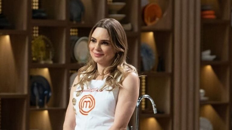 Mulher de cabelos loiros ondulados veste avental branco com o logotipo do MasterChef e nome bordado em vermelho, sorrindo levemente em um ambiente de estúdio culinário com prateleiras de madeira ao fundo exibindo pratos decorativos e iluminação suave.