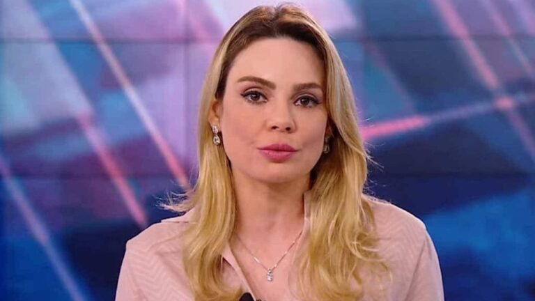 Mulher loira, de expressão séria, aparece em close diante de um fundo iluminado com tons azulados e linhas geométricas, como se estivesse apresentando um telejornal.