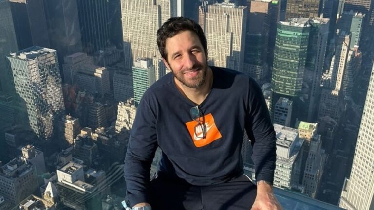 Homem de barba e blusa azul marinho com logotipo alaranjado posa sorrindo em mirante com vista panorâmica de arranha-céus ao fundo