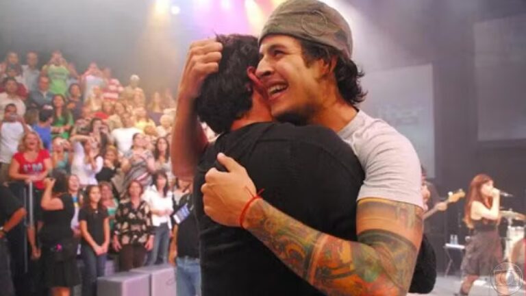 Homem sorridente com tatuagens e gorro abraça outro homem em palco de programa de TV com plateia e banda ao fundo