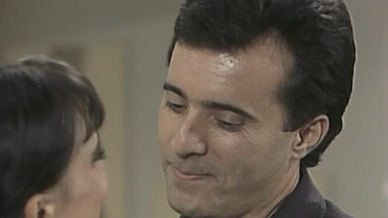 Close em um homem olhando de forma suave para uma mulher que aparece parcialmente de costas. Ambos estão em um ambiente interno, e a cena transmite um momento de intimidade e conversa próxima.