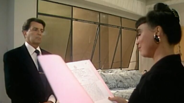 Cena interna com duas pessoas em ambiente formal. À esquerda, um homem de terno escuro e gravata observa com expressão séria. À direita, uma mulher com cabelo preso em coque e brincos grandes lê um documento dentro de uma pasta rosa, vista de perfil. O cenário tem paredes claras e painéis de vidro ao fundo, sugerindo um escritório ou instituição oficial.