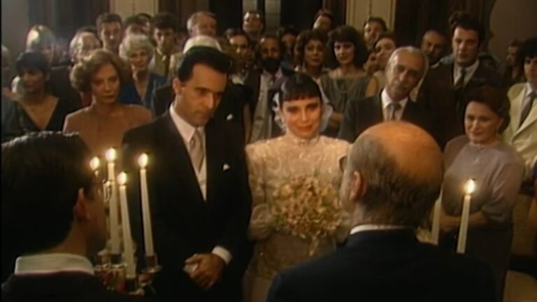Cena de casamento em ambiente interno, com iluminação quente. No centro, um casal de noivos está diante de um celebrante: o noivo usa terno escuro e a noiva veste um vestido claro de mangas longas, com véu curto e buquê nas mãos. Velas acesas aparecem em primeiro plano, criando clima solene. Ao fundo, convidados bem vestidos lotam o espaço, observando a cerimônia com expressões atentas e formais, sugerindo uma cena clássica de novela ou filme de época.