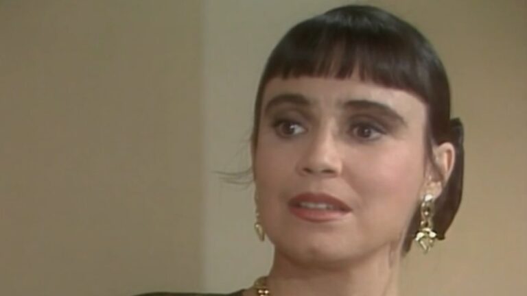 Mulher aparece em close, em ambiente interno de tom neutro, com expressão de surpresa ou apreensão, olhos bem abertos e boca levemente entreaberta. Ela tem cabelo escuro preso para trás, com franja reta curta, usa brincos dourados e colar delicado. A maquiagem é discreta, destacando os olhos. O enquadramento fechado e a expressão facial sugerem uma cena dramática, possivelmente de novela ou produção televisiva de época.