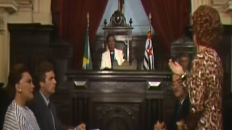 Cena em tribunal antigo com móveis de madeira escura mostra um juiz sentado ao centro vestindo paletó claro, atrás de bandeiras do Brasil e de São Paulo, enquanto uma mulher em pé fala gesticulando diante de três pessoas sentadas, duas mulheres e um homem, em ambiente com cortinas vermelhas e arquitetura clássica