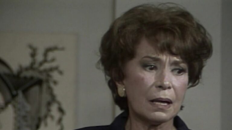 Mulher mais velha, de expressão aflita e preocupada, olha para o lado em um ambiente interno. Ela usa brincos dourados e tem o cabelo curto e castanho. A iluminação suave destaca suas rugas de tensão, sugerindo um momento emocional delicado