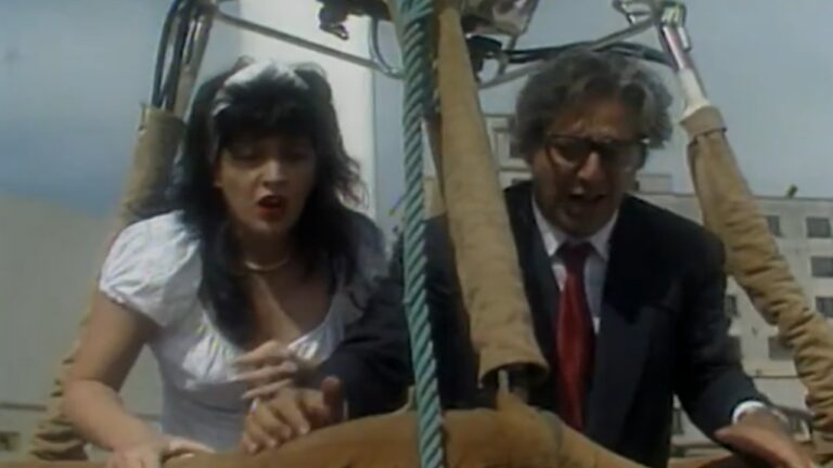 Um homem e uma mulher aparecem em um cesto de balão de ar quente, inclinados para a frente e com expressões de aflição. A mulher, de vestido branco e cabelo escuro, segura a borda do cesto, enquanto o homem, de terno escuro e gravata vermelha, também se apoia, aparentando tensão. Ao fundo, veem-se prédios sob um céu claro.