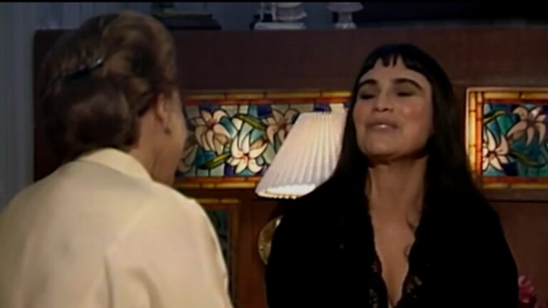 Duas mulheres adultas conversam em um ambiente interno com iluminação quente. À direita, uma mulher de cabelos longos e escuros, com franja reta, sorri levemente enquanto fala; ela veste roupa escura com decote discreto. À esquerda, outra mulher aparece de costas ou de perfil, com cabelo preso e roupa clara. Ao fundo, há um móvel de madeira, um abajur aceso e um painel decorativo em vitral, sugerindo uma cena doméstica ou dramática de produção televisiva.