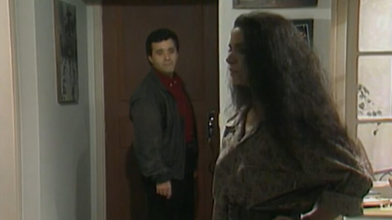 Cena em ambiente interno mostra um homem parado à porta de um apartamento, olhando para dentro, enquanto uma mulher aparece em primeiro plano, de perfil, com a mão apoiada na cintura. O homem veste camisa vermelha e jaqueta escura; a mulher usa blusa de mangas longas e tem cabelos longos e soltos. O espaço é iluminado por luz natural que entra por uma janela à direita, sugerindo um momento de tensão ou despedida dentro de um ambiente doméstico.