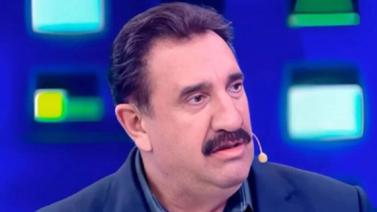 Homem adulto com bigode escuro participa de um programa de televisão, usando microfone de lapela preso ao rosto. Ele veste terno escuro e camisa clara e olha para o lado com expressão séria e atenta. Ao fundo, há um cenário de estúdio em tons de azul, com formas geométricas iluminadas, característico de um talk show ou programa jornalístico na TV.