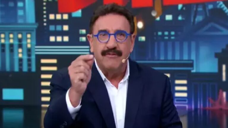 Homem de meia-idade aparece em estúdio de televisão, usando terno escuro, camisa branca e óculos de armação azul. Ele tem bigode e fala diretamente para a câmera, fazendo um gesto com a mão direita. Ao fundo, há um cenário gráfico com ilustrações estilizadas de prédios e luzes urbanas, em tons de azul e vermelho.