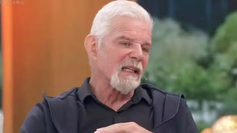 Homem idoso em estúdio de TV, visto do peito para cima, com cabelos brancos curtos e barba branca. Ele veste camisa preta e fala enquanto gesticula levemente, com expressão séria e reflexiva. O fundo é desfocado, com tons quentes e elementos que lembram plantas, indicando um ambiente de programa televisivo.