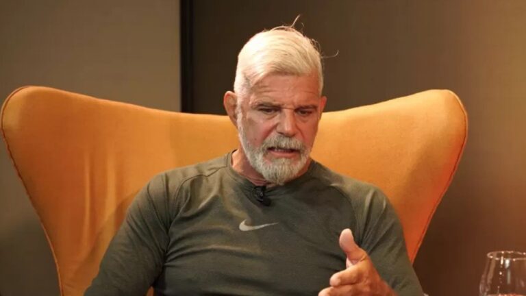 Homem de cabelos e barba grisalhos está sentado em uma poltrona laranja, falando e gesticulando com a mão. Ele veste camiseta esportiva verde-oliva com símbolo da Nike. A iluminação é quente e há uma taça de vidro parcialmente visível à frente