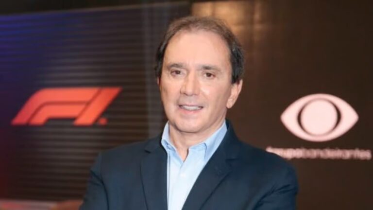 Homem de meia-idade, pele clara, usando terno escuro e camisa clara, posa de frente para a câmera com expressão neutra. Ao fundo, há um painel escuro com logotipos iluminados, incluindo um símbolo vermelho à esquerda e um ícone em forma de olho à direita, sugerindo um evento corporativo ou de mídia. O ambiente é bem iluminado e tem aparência formal.