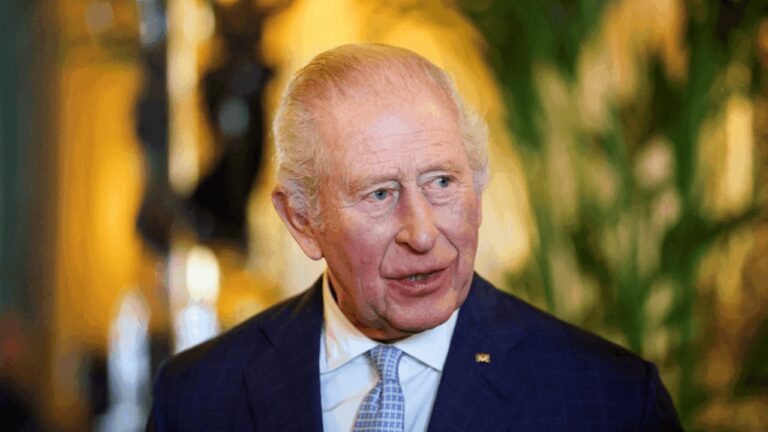 Homem mais velho, de cabelos grisalhos penteados para trás, veste terno azul escuro e gravata clara. Ele aparece em ambiente interno elegante, com iluminação quente e elementos decorativos desfocados ao fundo, enquanto olha levemente para o lado.