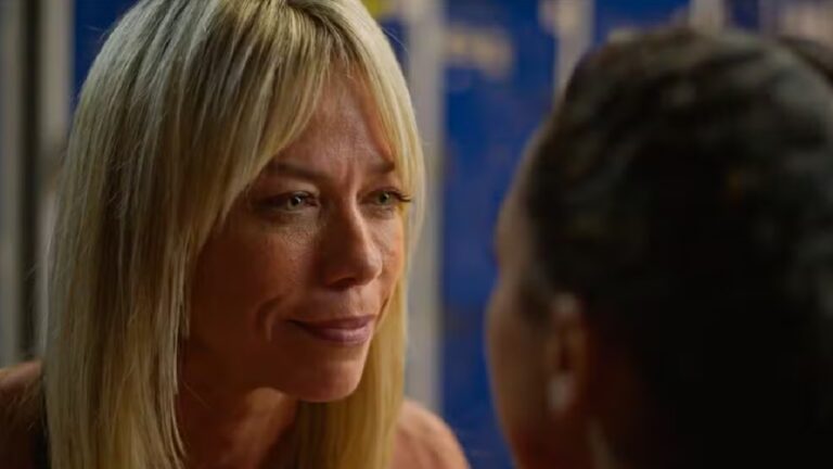 Close-up de uma mulher loira, de pele clara, com expressão emocionada e olhar atento, conversando de perto com outra pessoa que aparece de costas em primeiro plano. A mulher tem cabelos lisos e longos, maquiagem discreta e olhos levemente marejados, sugerindo um momento íntimo ou tenso. O fundo é desfocado, com tons de azul, destacando o clima dramático da cena.