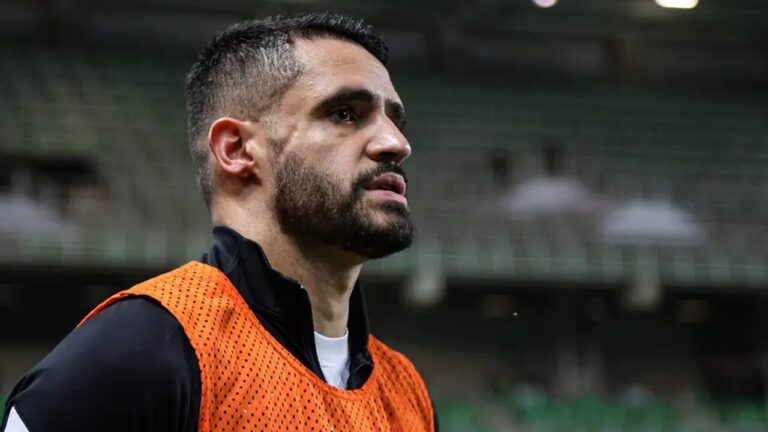 Jogador de futebol com barba e cabelo curto usa colete laranja durante treino em estádio vazio com arquibancadas ao fundo