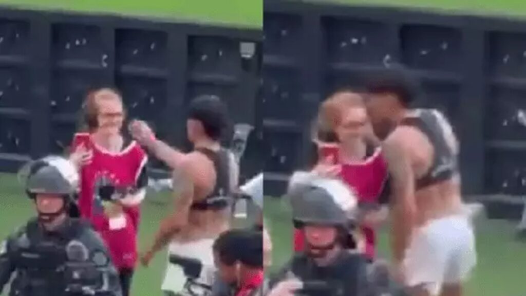 Duas cenas lado a lado mostram um jogador sem camisa, usando colete de monitoramento esportivo, aproximando-se de uma fotógrafa com colete vermelho à beira do campo. Ele estende a mão para o rosto dela e, na sequência, inclina-se para lhe dar um beijo na bochecha. Policiais equipados com capacetes aparecem no plano inferior, enquanto o fundo exibe arquibancadas escuras