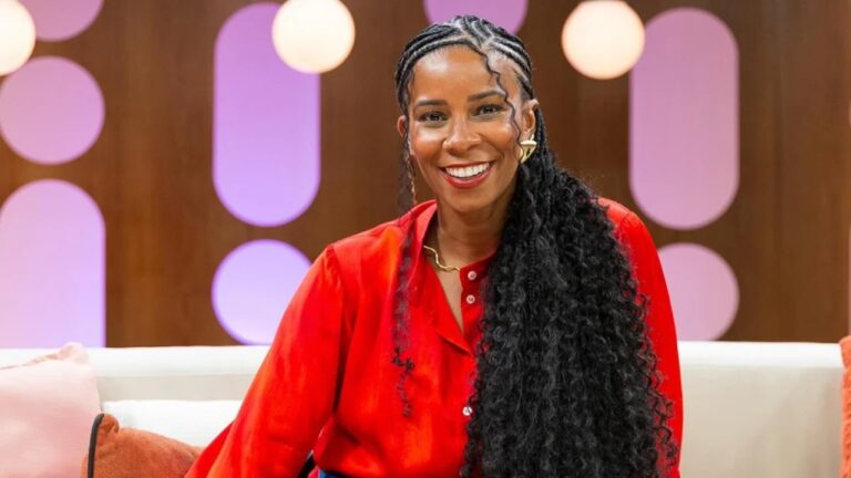 Mulher negra sorri sentada em sofá de estúdio vestindo blusa vermelha com cabelo trançado e iluminação decorativa ao fundo