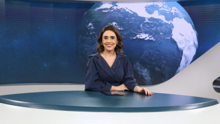 Jornalista sorridente sentada à bancada de telejornal com imagem da Terra ao fundo no estúdio de notícias