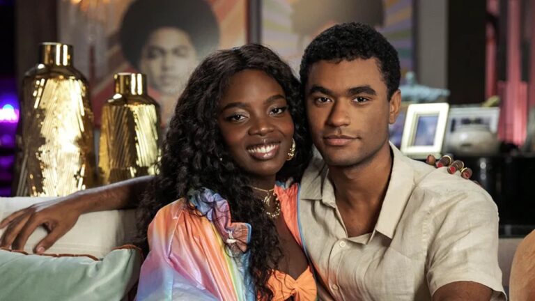 Casal jovem sentado em um sofá, posando lado a lado e olhando para a câmera. A mulher, de pele negra e cabelo longo e cacheado, sorri e usa um vestido colorido com brincos dourados; o homem, de pele morena e cabelo curto, veste camisa clara e mantém expressão serena. O ambiente é interno e decorado, com vasos dourados, quadros ao fundo e iluminação aconchegante, sugerindo uma cena de novela ou produção televisiva.