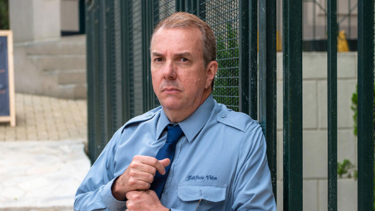 Augusto Madeira interpreta Rivaldo de uniforme azul e gravata encostado na grade verde em cena da novela Três Graças