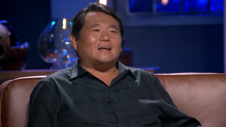 Homem sentado em uma poltrona de couro, sorrindo enquanto participa de uma entrevista em estúdio. Ele usa camisa preta e está em destaque no centro da imagem. O fundo é desfocado, com iluminação azulada e objetos decorativos pouco nítidos