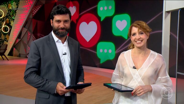 A imagem mostra Rodrigo Carvalho e Poliana Abritta no estúdio do programa Fantástico, da TV Globo. Eles estão sorrindo e segurando tablets, prontos para introduzir uma reportagem. O cenário tem tons vermelhos e dourados, com ícones digitais de corações e balões de mensagem exibidos em um telão ao fundo, sugerindo uma pauta sobre redes sociais ou relacionamentos