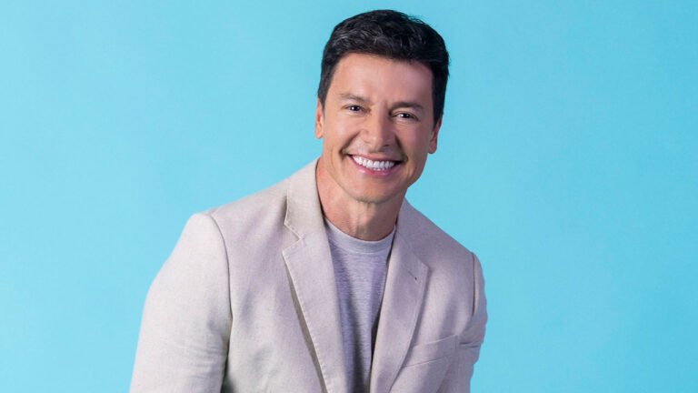 Rodrigo Faro de blazer bege e camiseta cinza sorri para a câmera em foto promocional com fundo azul