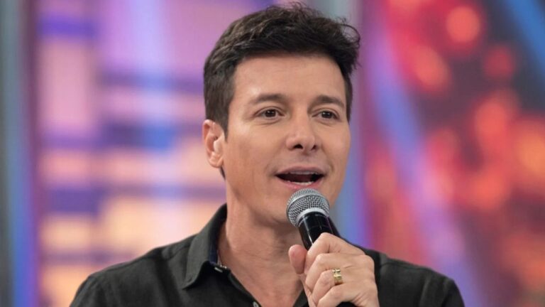 Apresentador segurando um microfone enquanto fala, enquadrado em close. Ele veste camisa escura e aparece diante de um fundo desfocado com luzes coloridas, sugerindo um estúdio de TV.