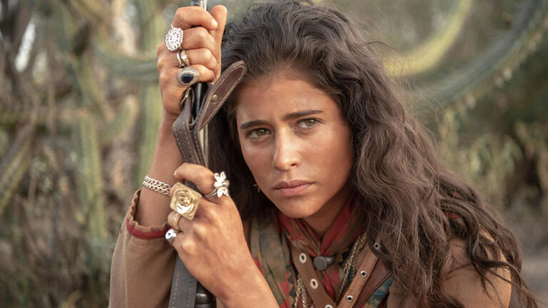 Isadora Cruz como Rosa com cabelos escuros e ondulados usando muitos anéis e segurando uma espingarda em Guerreiros do Sol
