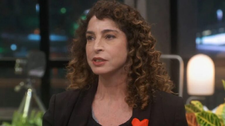 Mulher de cabelos castanhos encaracolados, usando blazer preto com um broche vermelho em formato de coração, aparece falando em um estúdio iluminado. Ao fundo, luzes desfocadas e elementos de decoração