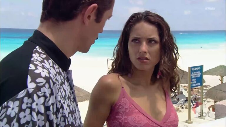 A imagem mostra uma cena de Rubi, novela gravada em uma praia de águas azul-turquesa. Uma mulher com expressão séria ou desconfiada, vestindo um vestido rosa, olha para um homem que está de costas parcialmente viradas para a câmera. Ele usa uma camisa com estampa floral preta e branca. Ao fundo, há guarda-sóis e pessoas na areia, além de uma placa de aviso em inglês. O logotipo no canto inferior direito indica que a cena foi exibida pelo SBT