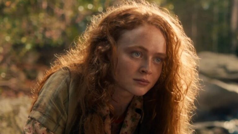 Jovem com cabelos ruivos e soltos observa atentamente para frente em ambiente externo com árvores e luz natural ao fundo