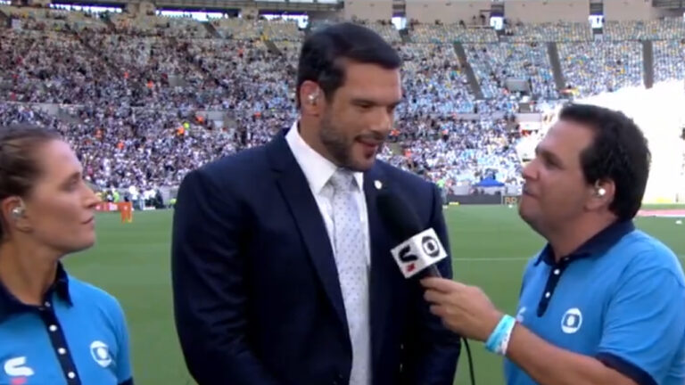 Samir Xaud de terno no gramado de estádio lotado ao lado de profissionais da Globo durante cobertura