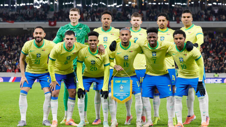 Jogadores da Seleção Brasileira de futebol posando para foto em campo, com uniforme amarelo e azul, segurando uma flâmula