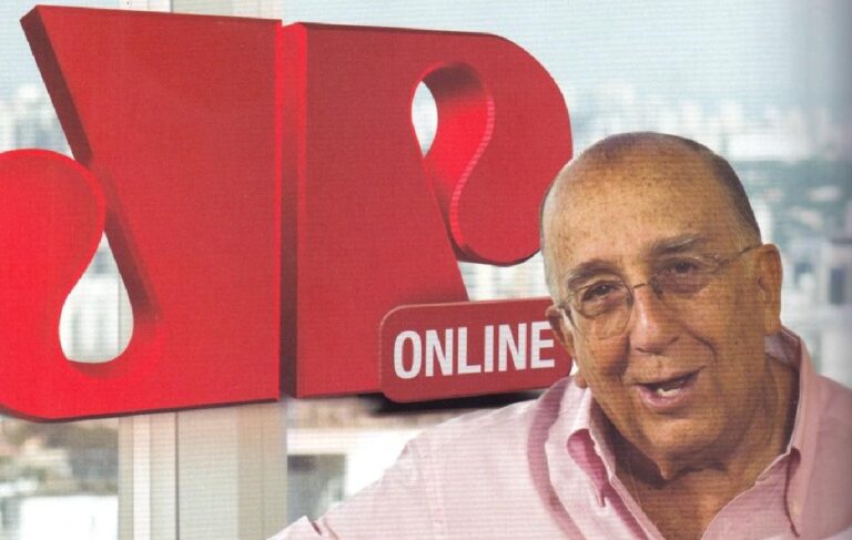 Homem mais velho, sorrindo, usando camisa rosa e óculos, posando diante de uma grande logomarca vermelha com as letras estilizadas “JP” e o termo “ONLINE”. Ao fundo, vê-se uma paisagem urbana desfocada através de janelas amplas