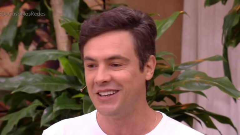 Homem sorridente, de cabelos castanhos curtos, veste camiseta clara e usa microfone de lapela no rosto. Ele aparece em ambiente interno decorado com plantas ao fundo