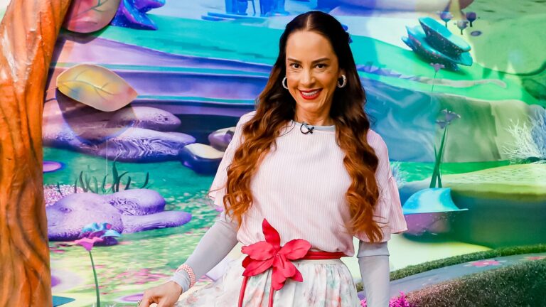 A imagem mostra Silvia Abravanel, uma mulher sorridente em um cenário colorido e lúdico, com elementos que lembram um jardim ou mundo encantado. Ela usa uma roupa clara com detalhes rosa, incluindo uma faixa na cintura com um laço em forma de flor. O ambiente e o figurino transmitem uma atmosfera alegre e voltada ao público infantil, em um programa exibido pelo SBT