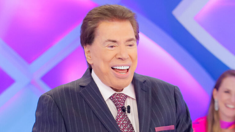 Silvio Santos sorrindo e olhando para a direita, de terno listrado e gravata vermelha, em um estúdio com luz rosa e azul