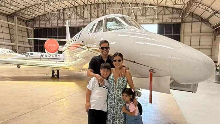 Um grupo de quatro pessoas posa sorrindo dentro de um hangar de aeronaves, em frente a um jato executivo branco. Um homem adulto está atrás, com as mãos apoiadas nos ombros de um menino à frente. Ao lado, uma mulher adulta usa vestido estampado e óculos escuros, abraçada a uma menina menor. Todos estão próximos à aeronave, que ocupa grande parte do fundo da imagem. O hangar é amplo, com estrutura metálica no teto e iluminação natural.