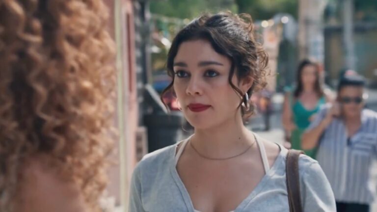 Mulher jovem olha atentamente para outra pessoa em uma rua movimentada durante o dia. Ela tem cabelos escuros presos de forma solta, usa brincos de argola, maquiagem discreta com batom vermelho e veste blusa clara com alça de bolsa no ombro. Ao fundo, pedestres e construções urbanas aparecem desfocados, indicando um ambiente cotidiano e urbano.