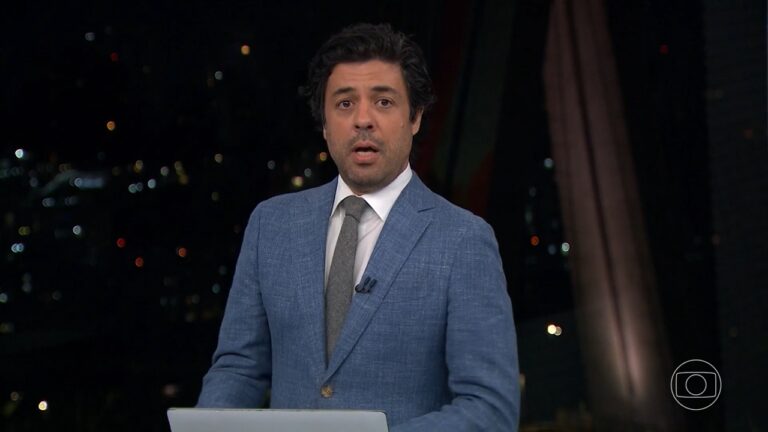 A imagem mostra Tiago Eltz, apresentador do telejornal SP2, em estúdio, usando um terno azul, camisa branca e gravata cinza. Ele está diante de um fundo escuro com luzes de cidade ao fundo, sugerindo uma transmissão noturna. À frente, há um laptop e, no canto inferior direito, aparece o logotipo da TV Globo