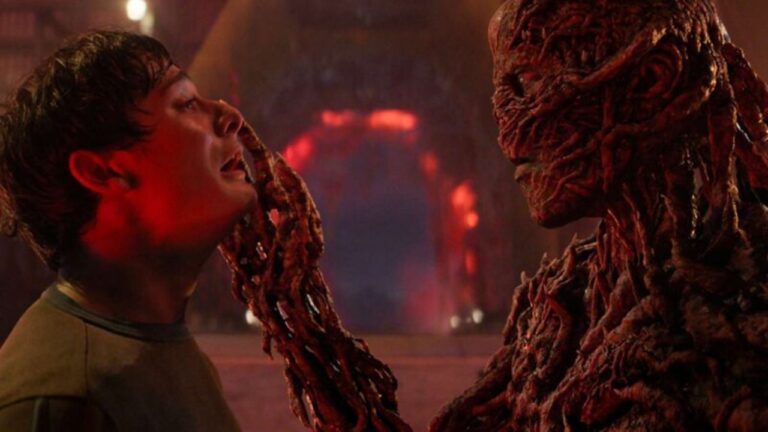 Cena com iluminação vermelha intensa mostra um jovem sendo segurado pelo rosto por uma criatura humanoide coberta por estruturas orgânicas e enrugadas, em ambiente sombrio com aparência de caverna iluminada por luzes alaranjadas ao fundo