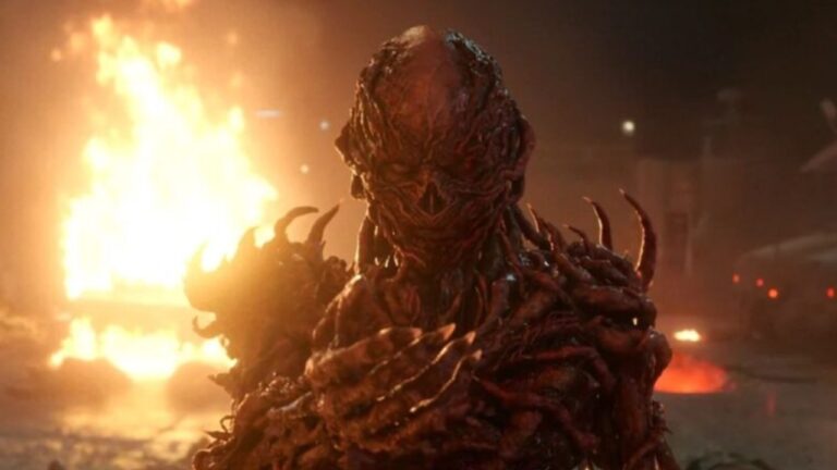 Criatura monstruosa de aparência humanoide, com pele avermelhada e textura orgânica semelhante a raízes retorcidas, aparece em primeiro plano com garras alongadas. Ao fundo, há um incêndio intenso iluminando a cena noturna, criando um clima sombrio e ameaçador